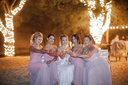 Bride and bridesmaids having a toast together in front of twinkle light wrapped trees